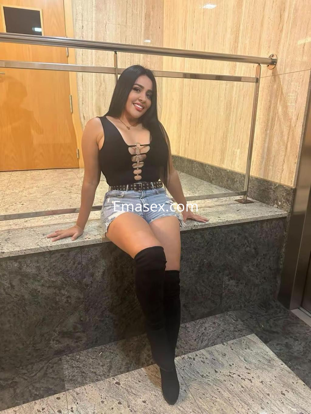 Escort en Santander, luciendo un top negro con detalles en el pecho y shorts de mezclilla, sentada en un entorno elegante.