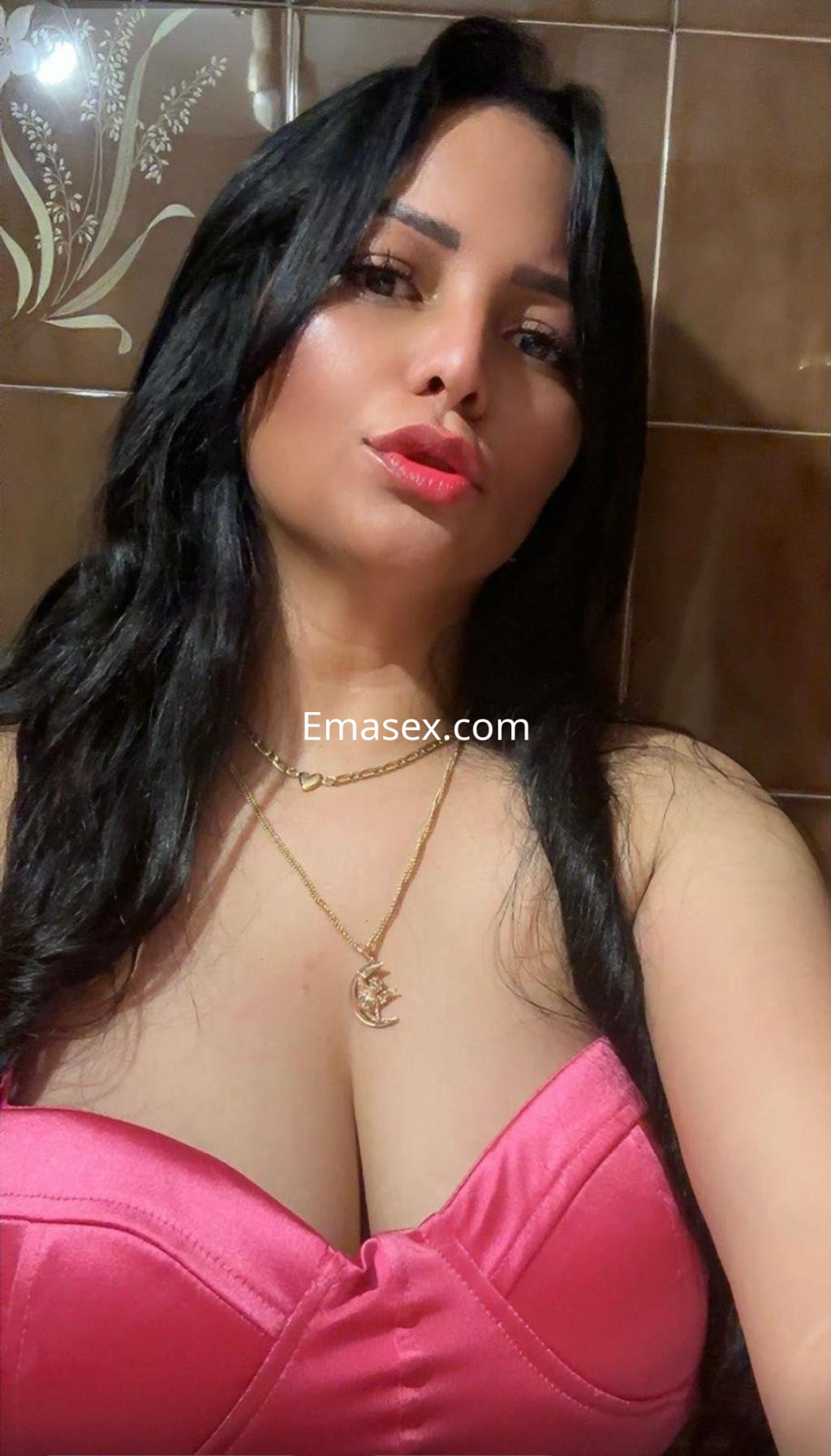 La puta posa con un vestido rosa, luciendo un collar dorado y maquillaje atractivo en un baño de Pamplona.