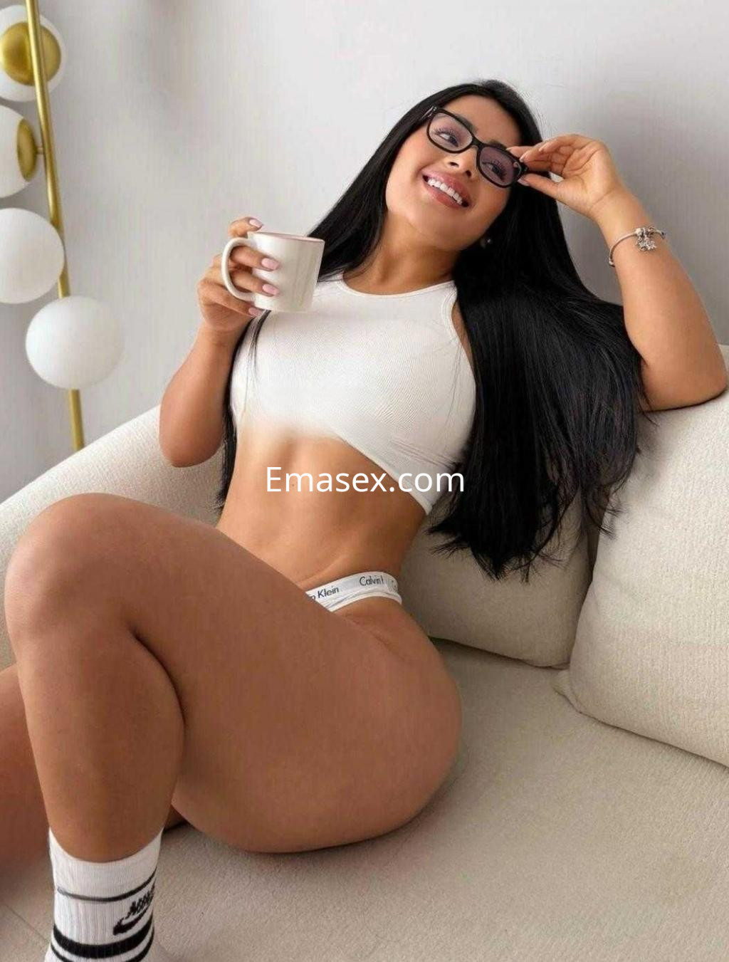 Escort en Barcelona sentada en un sofá, luciendo una blusa blanca y lencería, sosteniendo una taza sonriente.