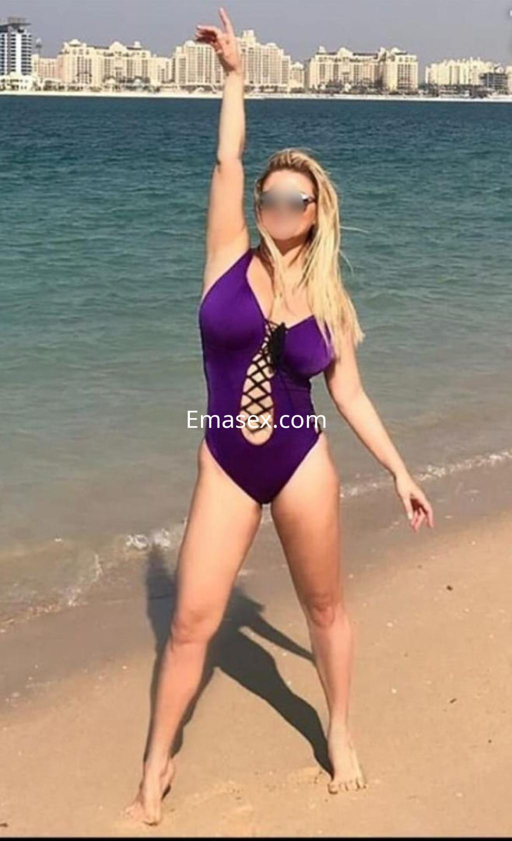 Puta en Granada luciendo un traje de baño morado en la playa, con el mar y edificios de fondo.