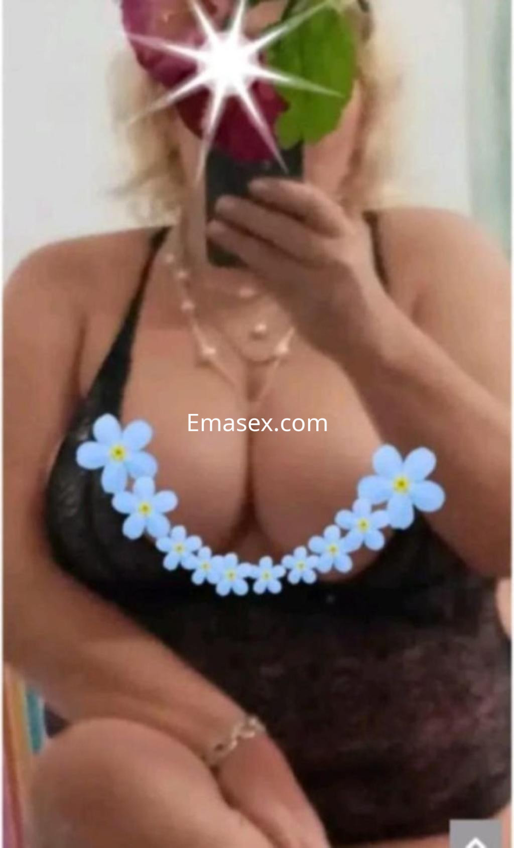 Puta en Murcia se muestra con lencería negra y un collar, mientras posa frente a un espejo con flores decorativas.