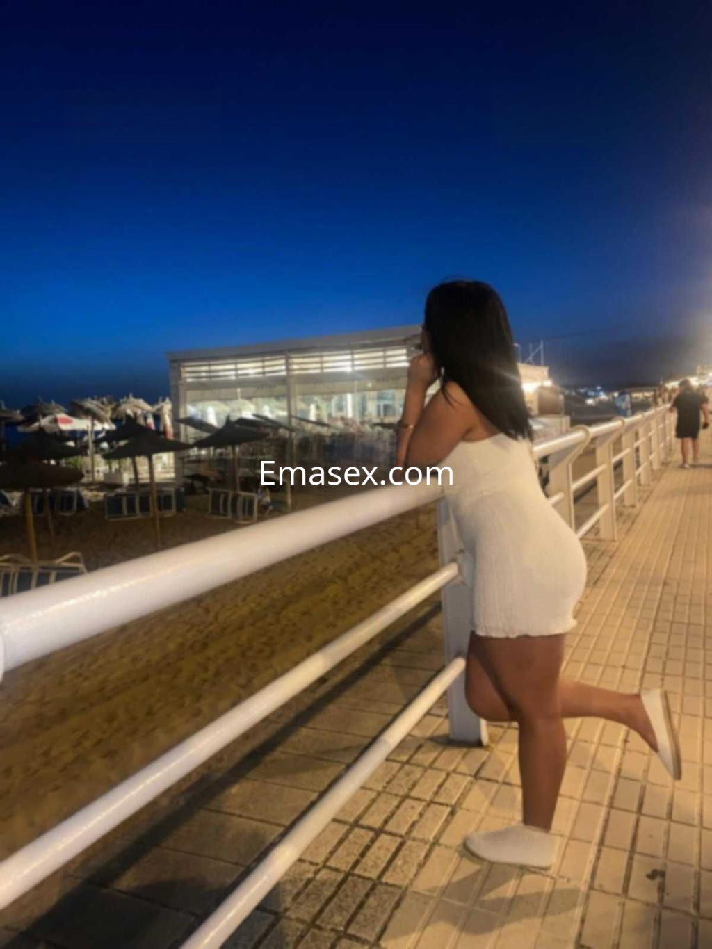 Escort en Sevilla apoyada en una barandilla, vistiendo un vestido blanco y con el mar al fondo al atardecer.