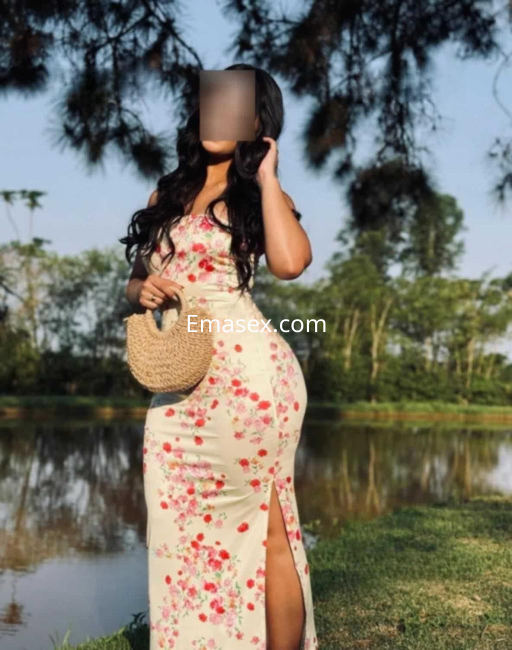 Puta en Huelva posando de pie junto a un lago, luciendo un vestido floral con una abertura y un bolso de mimbre.