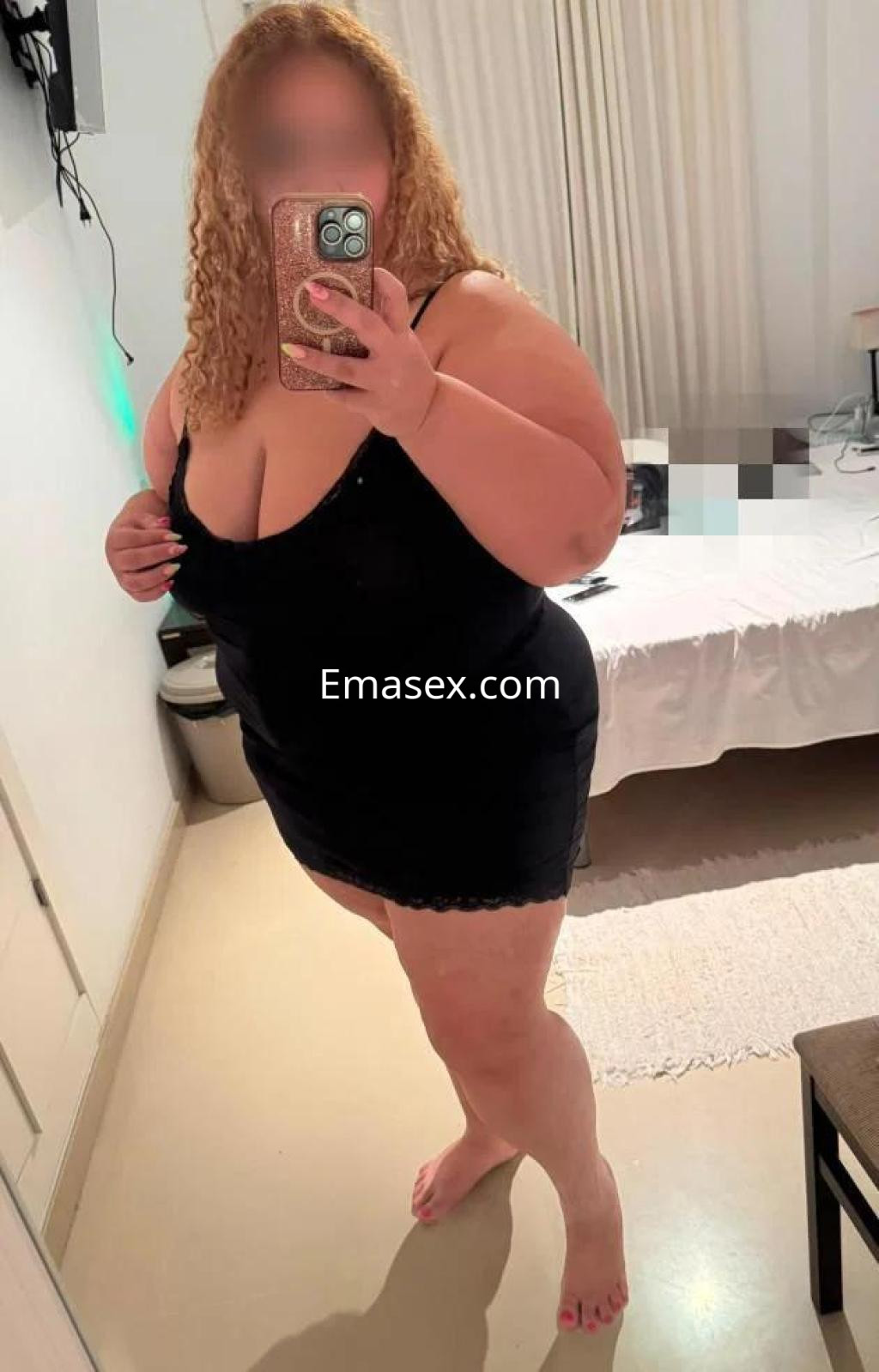 Escort en Logroño posando frente al espejo con un vestido negro ajustado y cabello rizado, en un dormitorio.