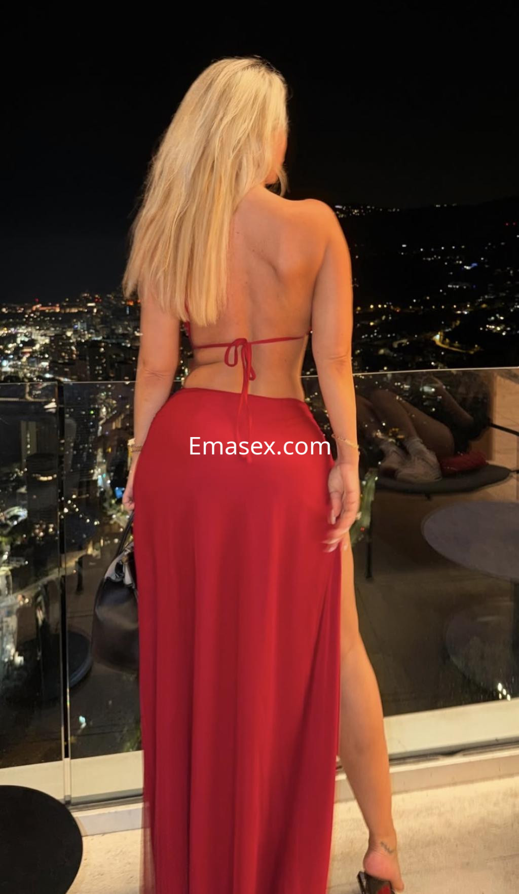 Escort de Tenerife con cabello rubio y largo, vistiendo un conjunto rojo y posando de espaldas frente a un paisaje urbano nocturno.