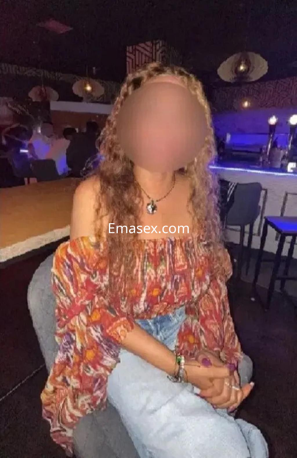 Escort en Oviedo sentada en un bar, lleva una blusa colorida de mangas largas y jeans claros.