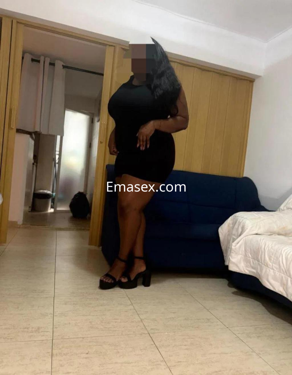 Puta de piel oscura con vestido negro ajustado y tacones, posando en una habitación en Valencia.