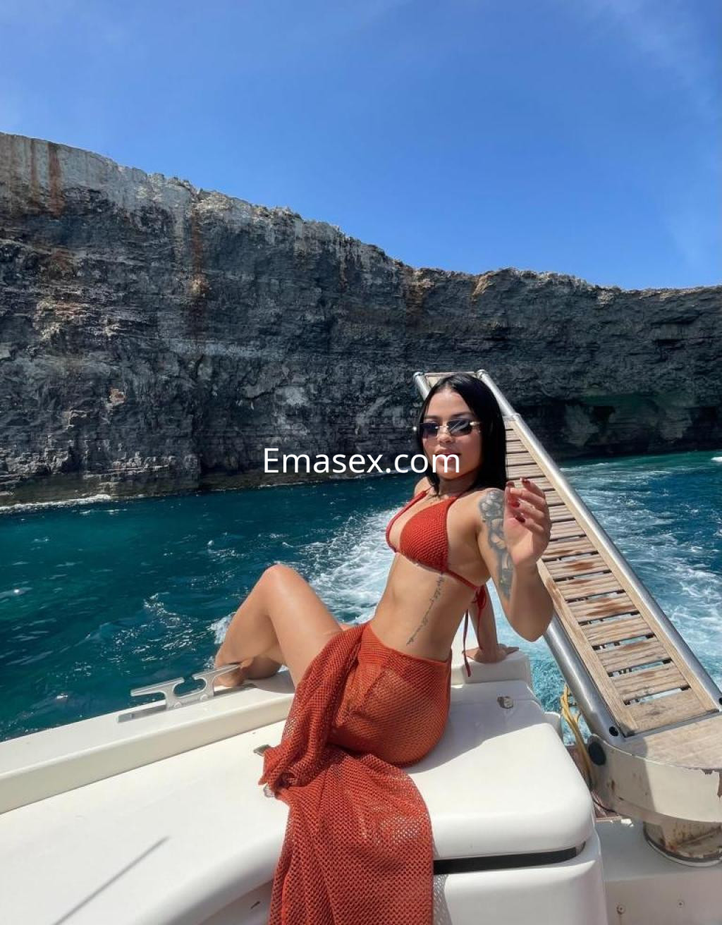 Escort en Málaga, posando en un barco con bikini rojo y pareo, con mar azul y acantilados al fondo.