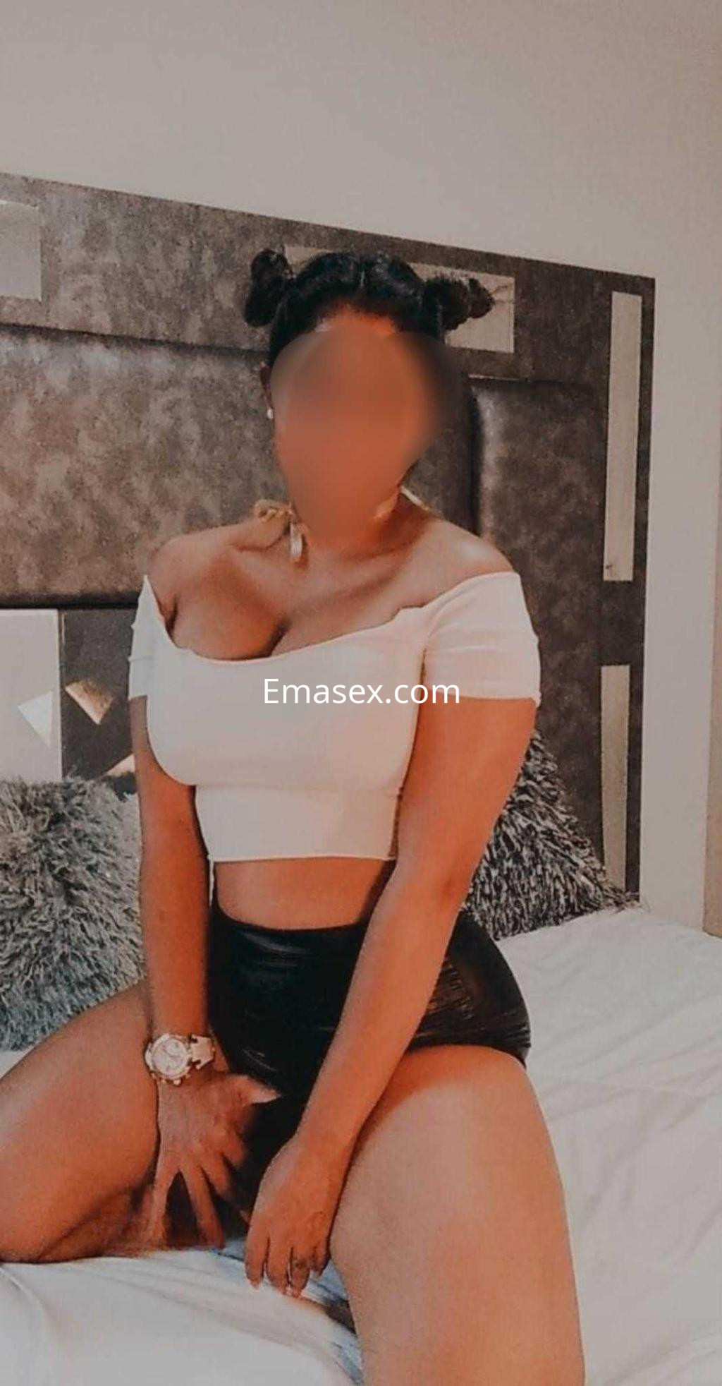 Puta en Málaga luciendo un top blanco y shorts negros, sentada en la cama con una pose sugestiva.