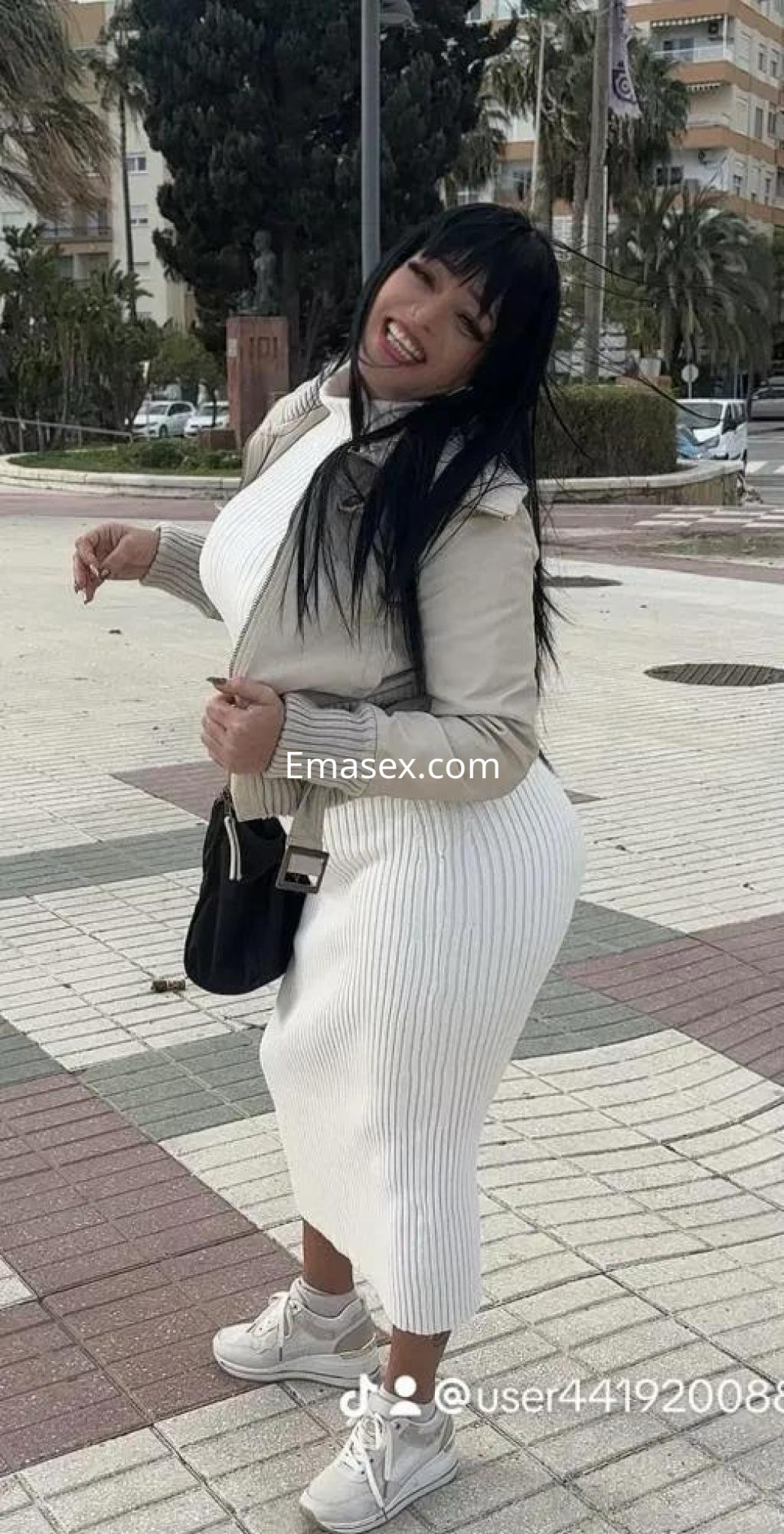Puta en Málaga, con cabello largo y oscuro, viste un vestido blanco y chaqueta beige, sonriendo en un entorno urbano.