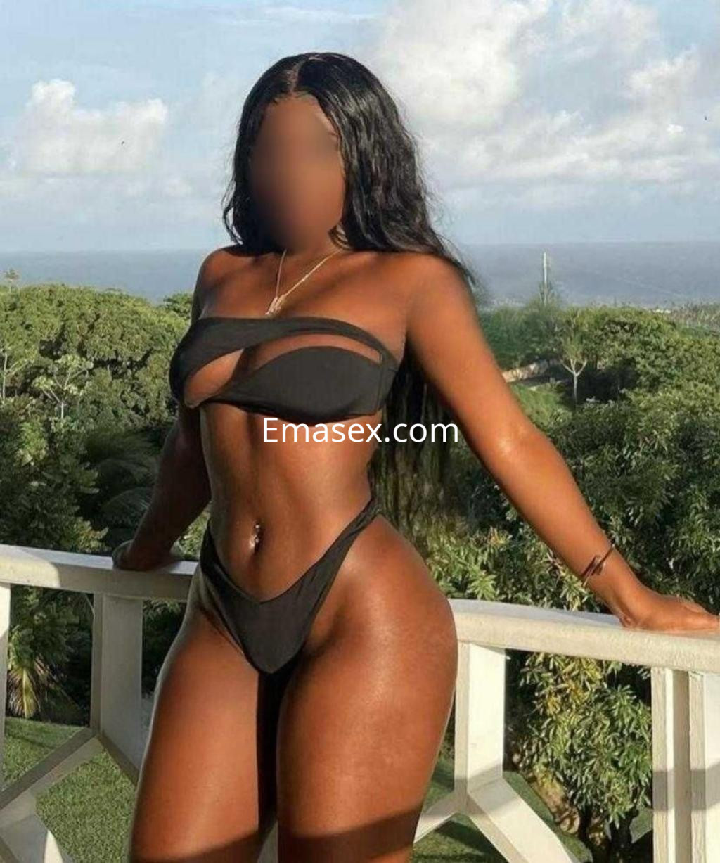 La puta se muestra de pie en un balcón, vistiendo un bikini negro que resalta su figura, con un paisaje tropical de fondo.