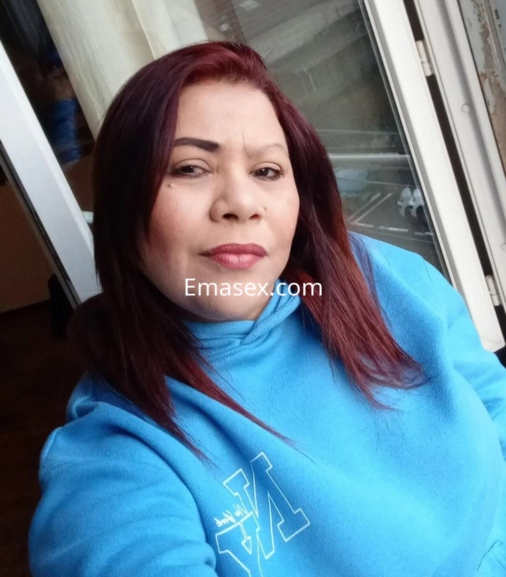 Escort en Zaragoza con cabello largo y lacio de color castaño, vistiendo sudadera azul y posando junto a una ventana.