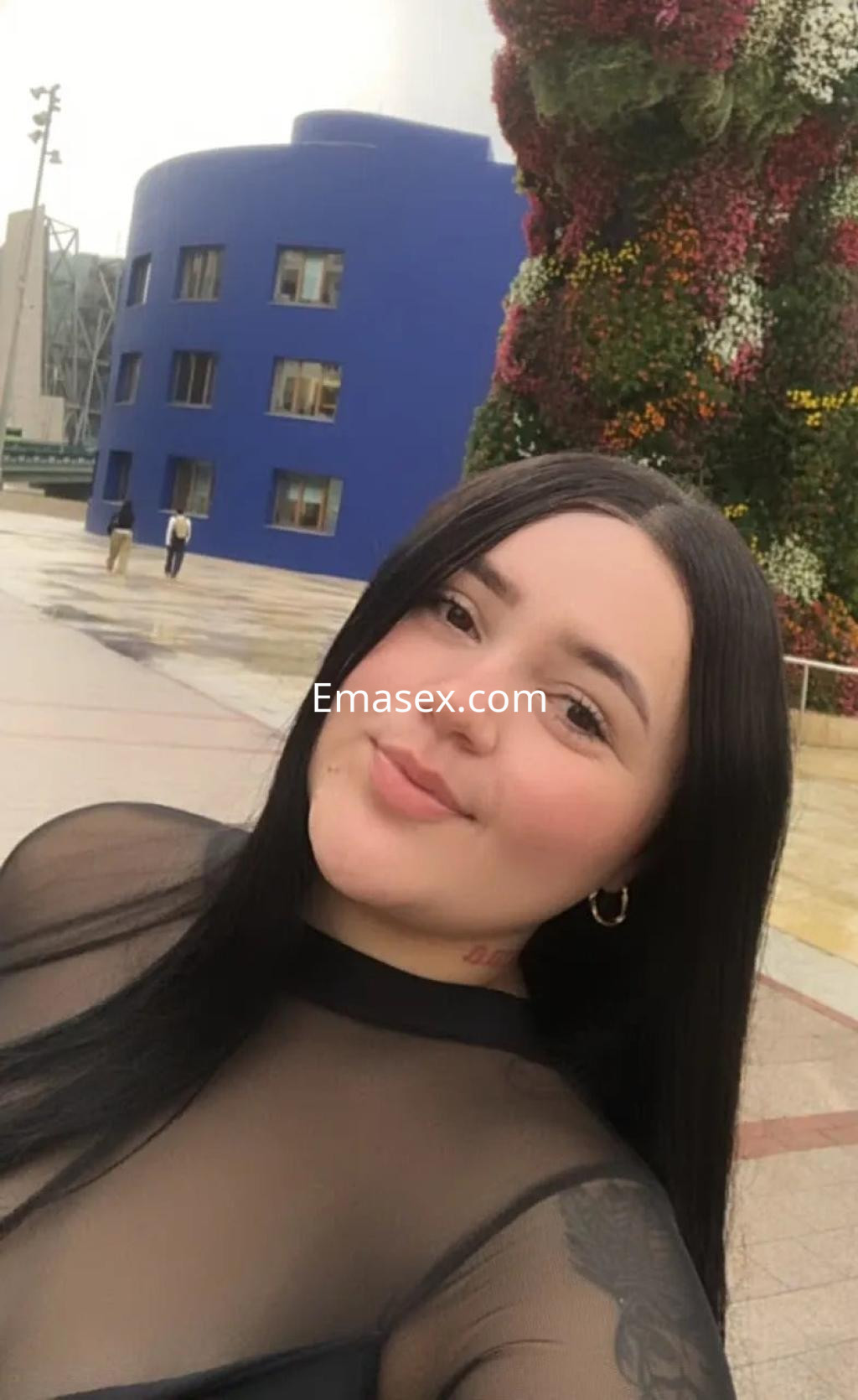 Escort en Oviedo sonríe para la cámara, vistiendo una blusa transparente y con el cabello largo y lacio, frente a un edificio azul.