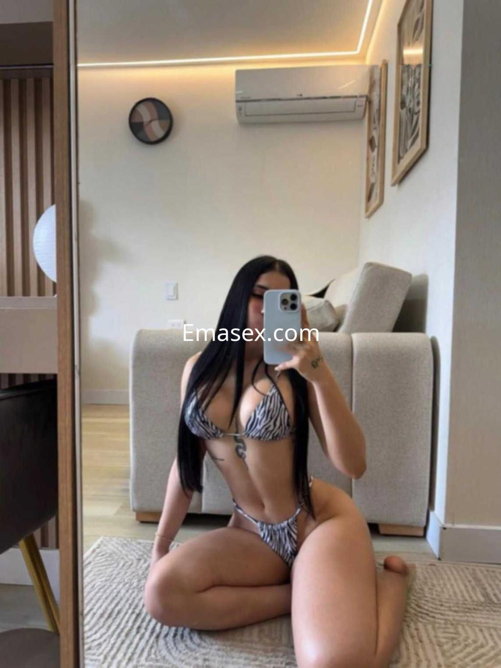 Escort en Tenerife posando en un espejo, viste un bikini de rayas y está sentada en el suelo de una sala moderna.