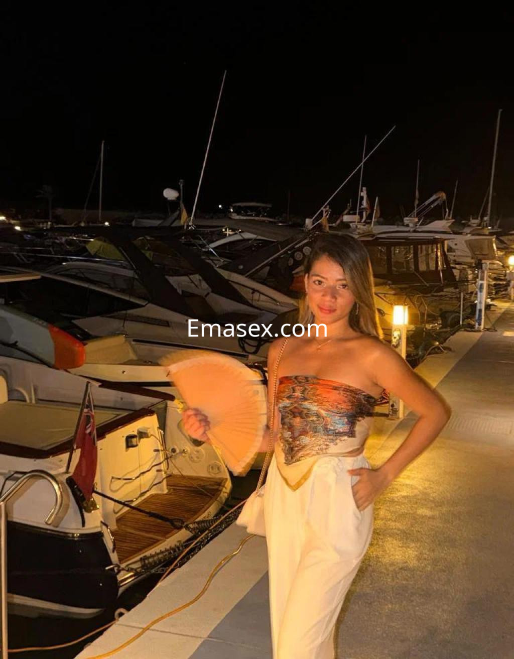 Escort en Palma de Mallorca, posando con un abanico, con un top sin mangas y pantalones claros, junto a yates.