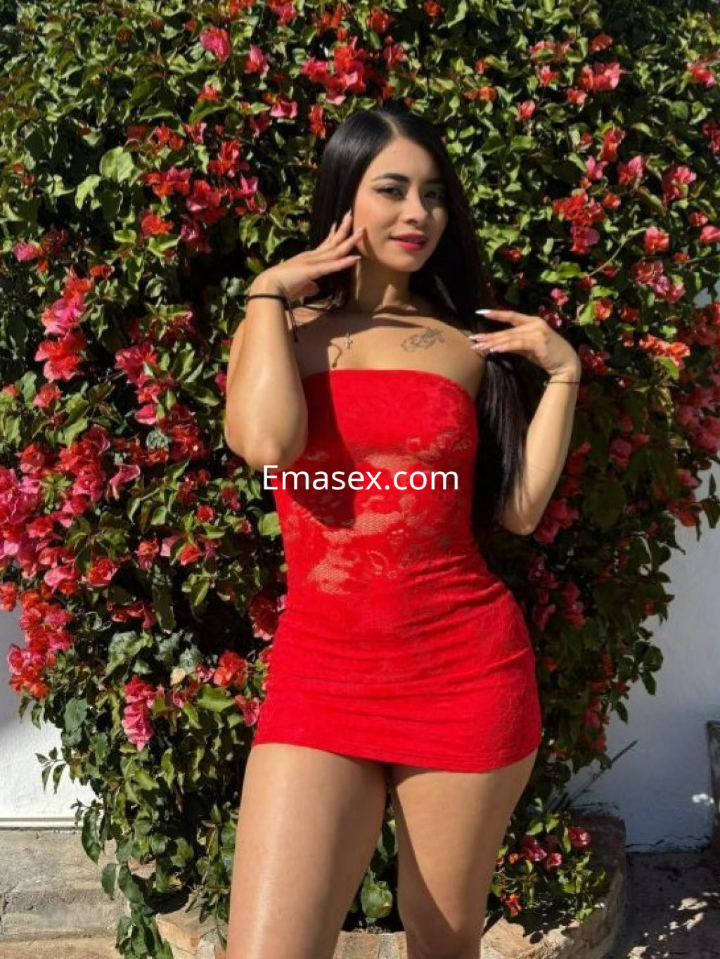Escort en Barcelona con cabello largo y lacio, viste un vestido rojo ajustado y posa entre flores coloridas.
