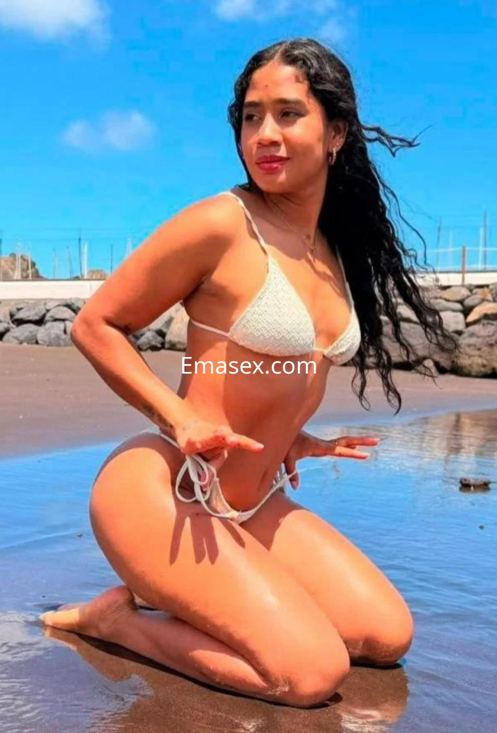 La escort en Almería se muestra de rodillas en la playa, luciendo un bikini blanco y el cabello ondulado suelto.