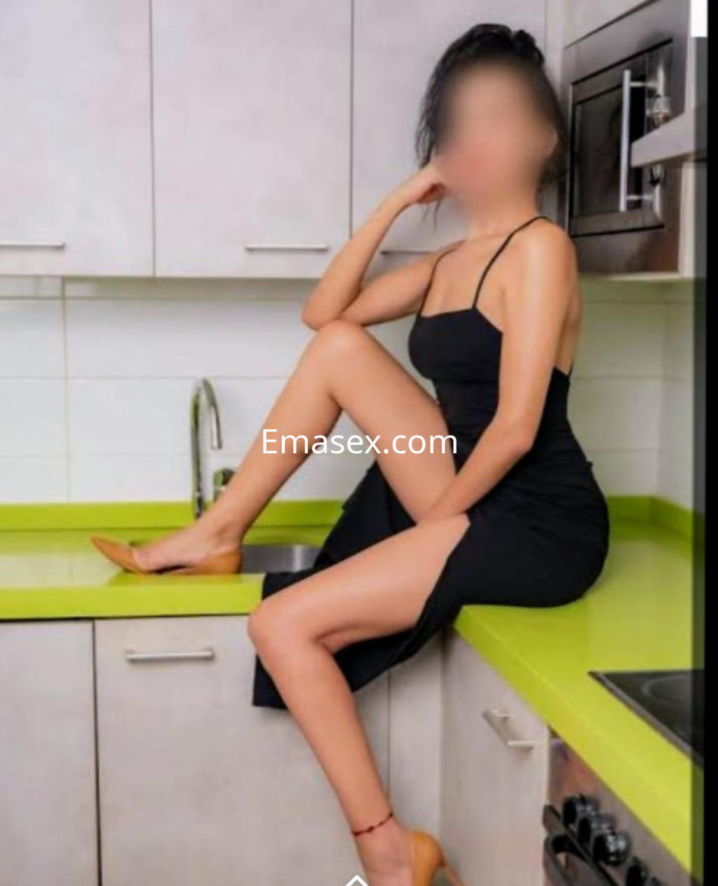 Puta sentada en una encimera de cocina, viste un vestido negro y tacones, con el pelo recogido y una pierna levantada.