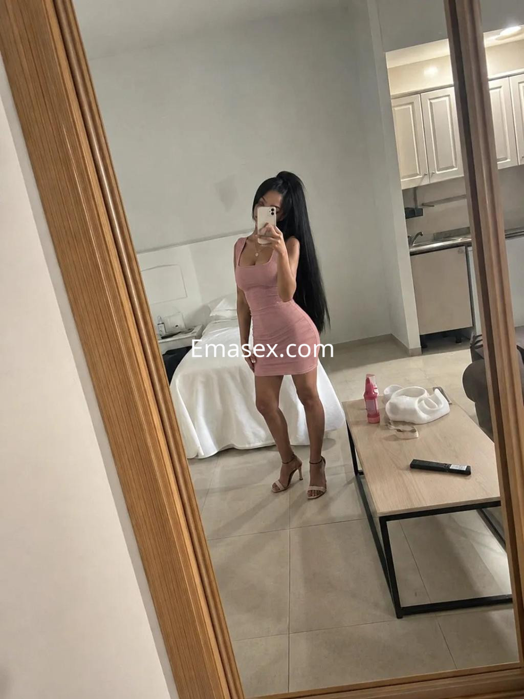 La puta en Málaga lleva un vestido rosa ajustado y sandalias, posando frente a un espejo en un ambiente moderno.