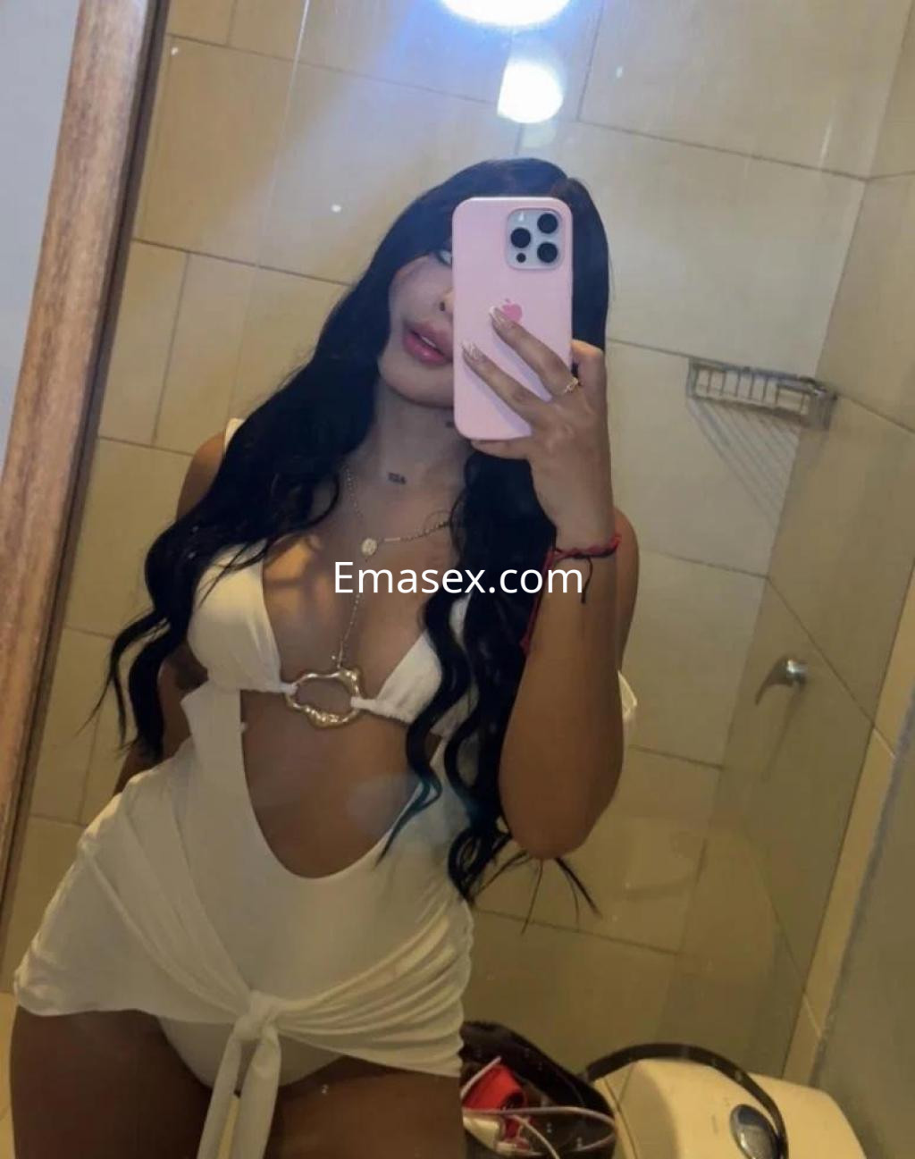 La puta en Tenerife posa frente al espejo con un vestido blanco que destaca su figura y cabello largo.