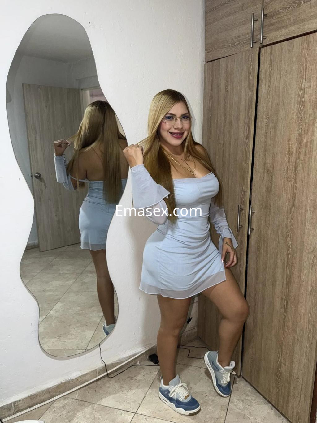 Escort en Sevilla posando junto a un espejo, luciendo un vestido ceñido de color claro y zapatillas deportivas.