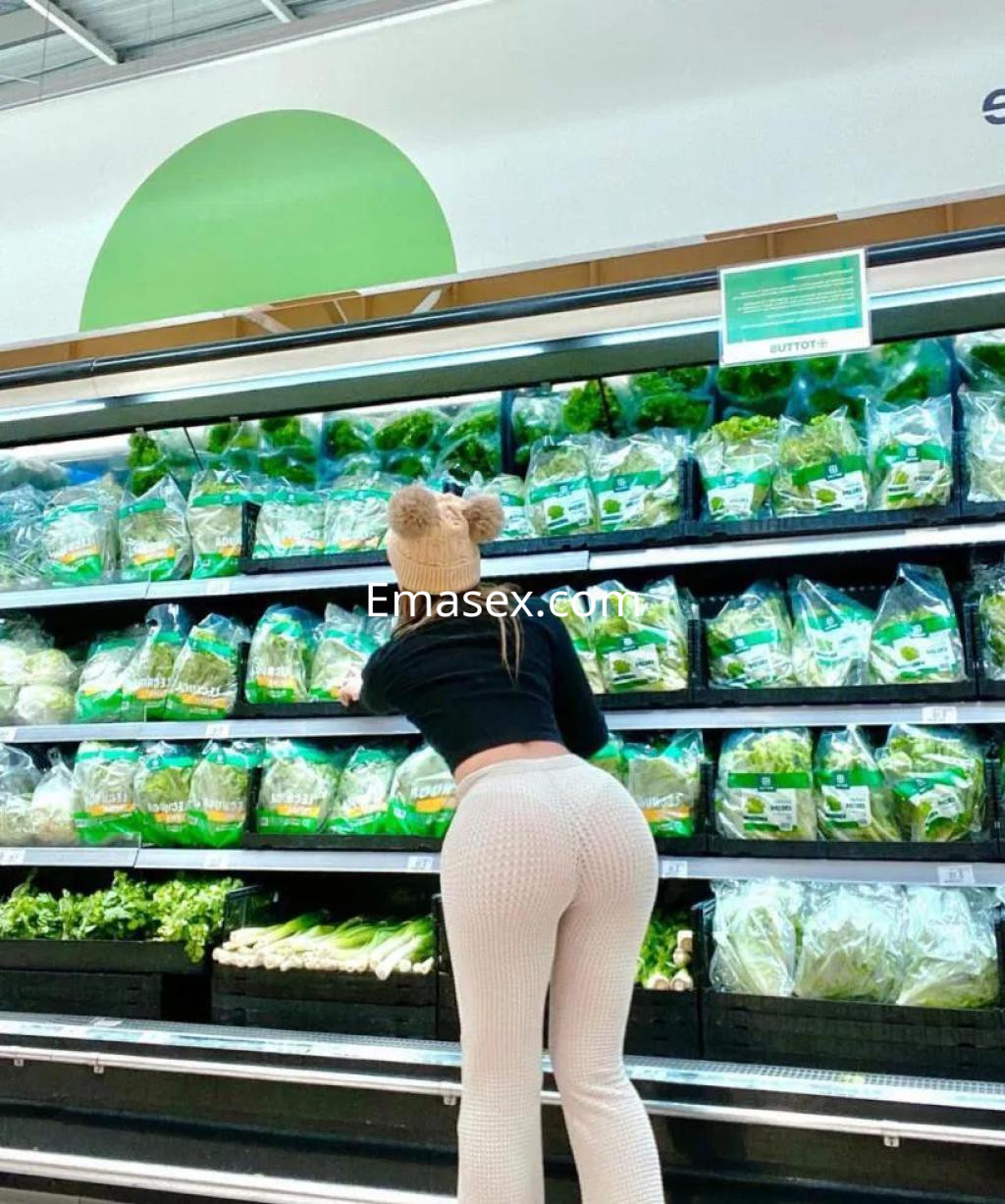 Puta en León vistiendo un conjunto negro y pantalones claros, agachada frente a estantes de verduras.