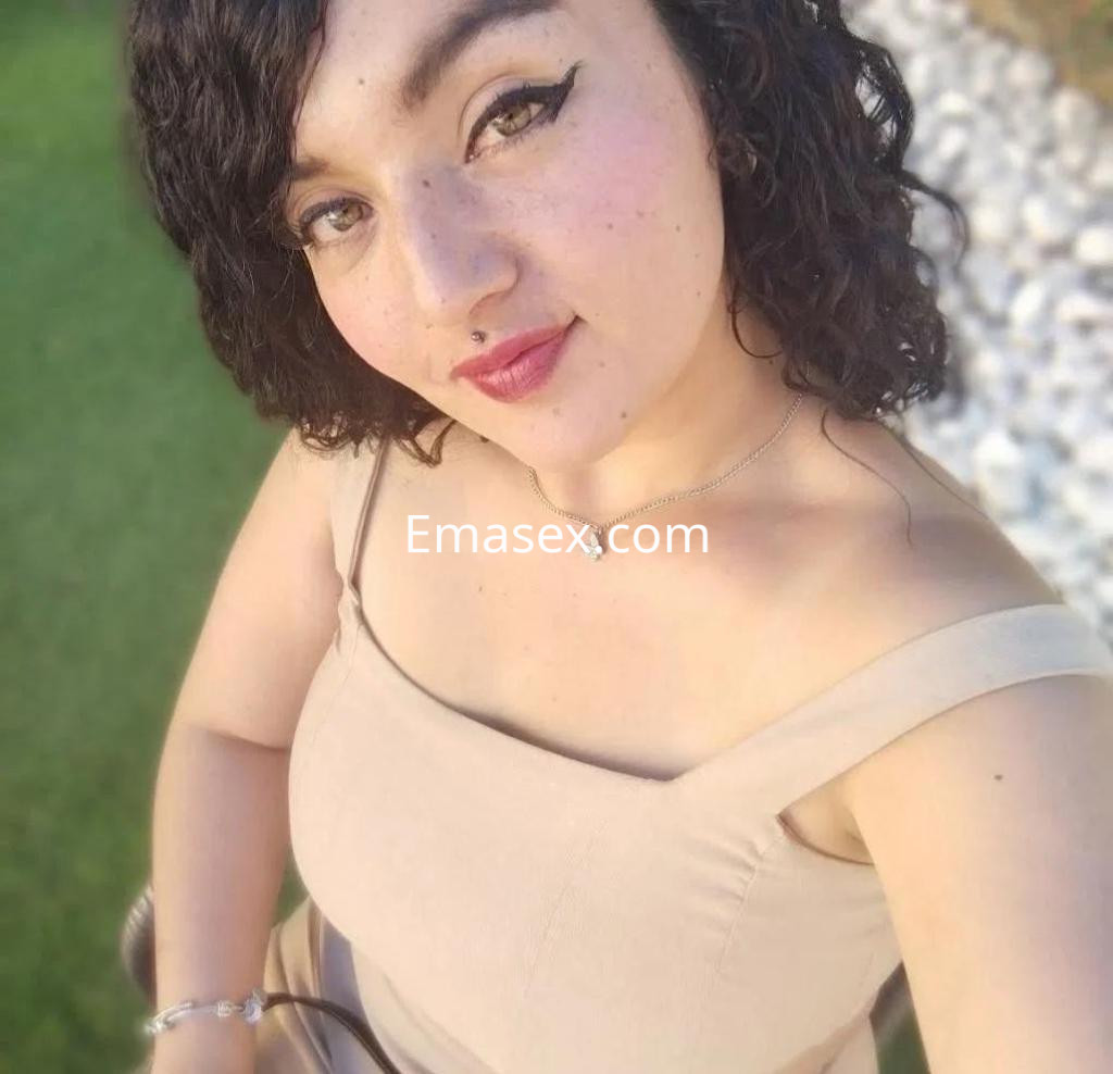 Puta con cabello rizado y oscuro, viste un vestido beige sin mangas, sonriente en un entorno verde en Lleida.