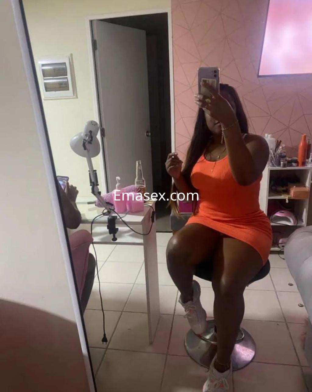 Puta en Lleida con vestido naranja ajustado, sentada en una silla, frente a un espejo.