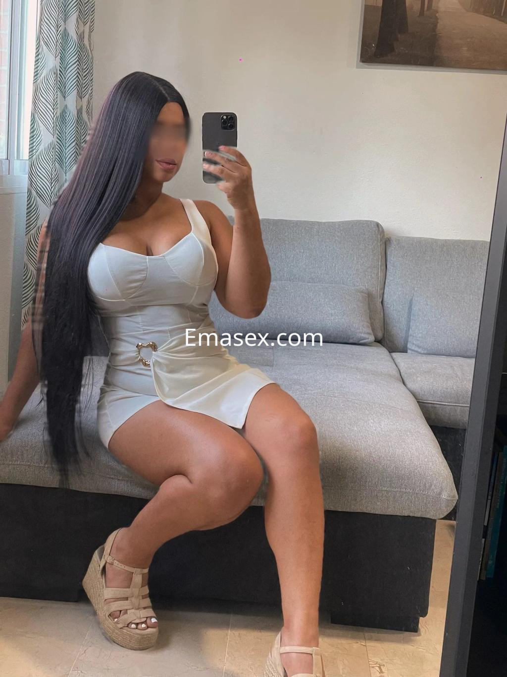 Puta con cabello largo y lacio, vistiendo un vestido blanco ajustado, sentada en un sofá en Sevilla.