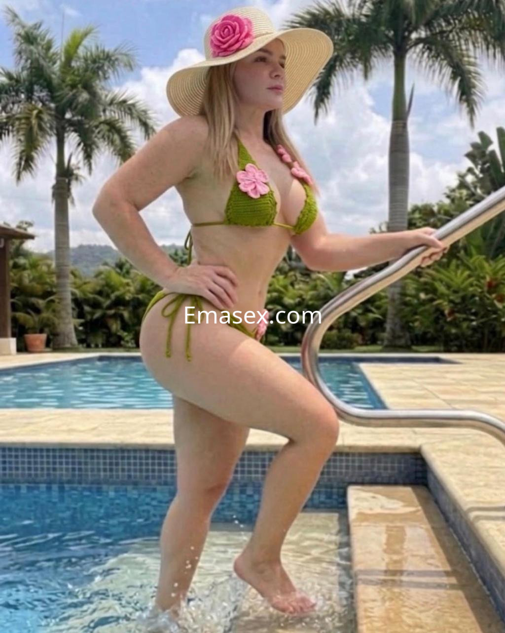 Escort en Pamplona con bikini verde y rosa, posando en una piscina bajo un sombrero grande y palm trees.
