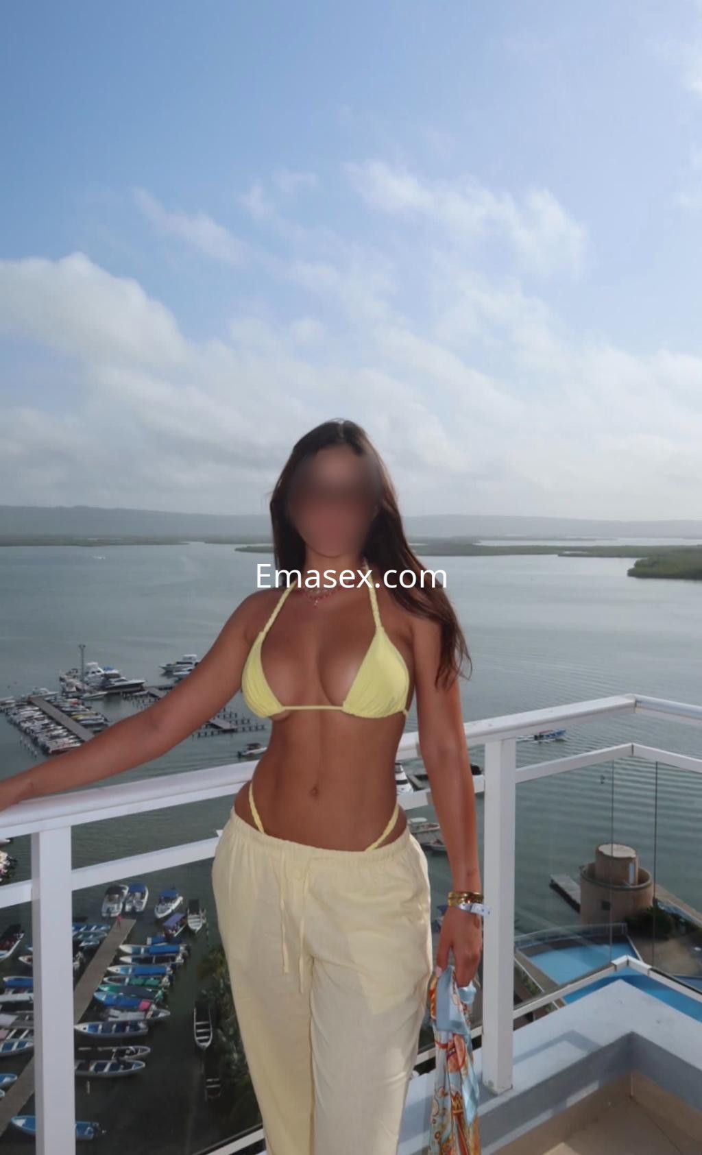 Escort en Bilbao, luciendo un bikini amarillo y pantalones claros, con vista al mar y un puerto de fondo.
