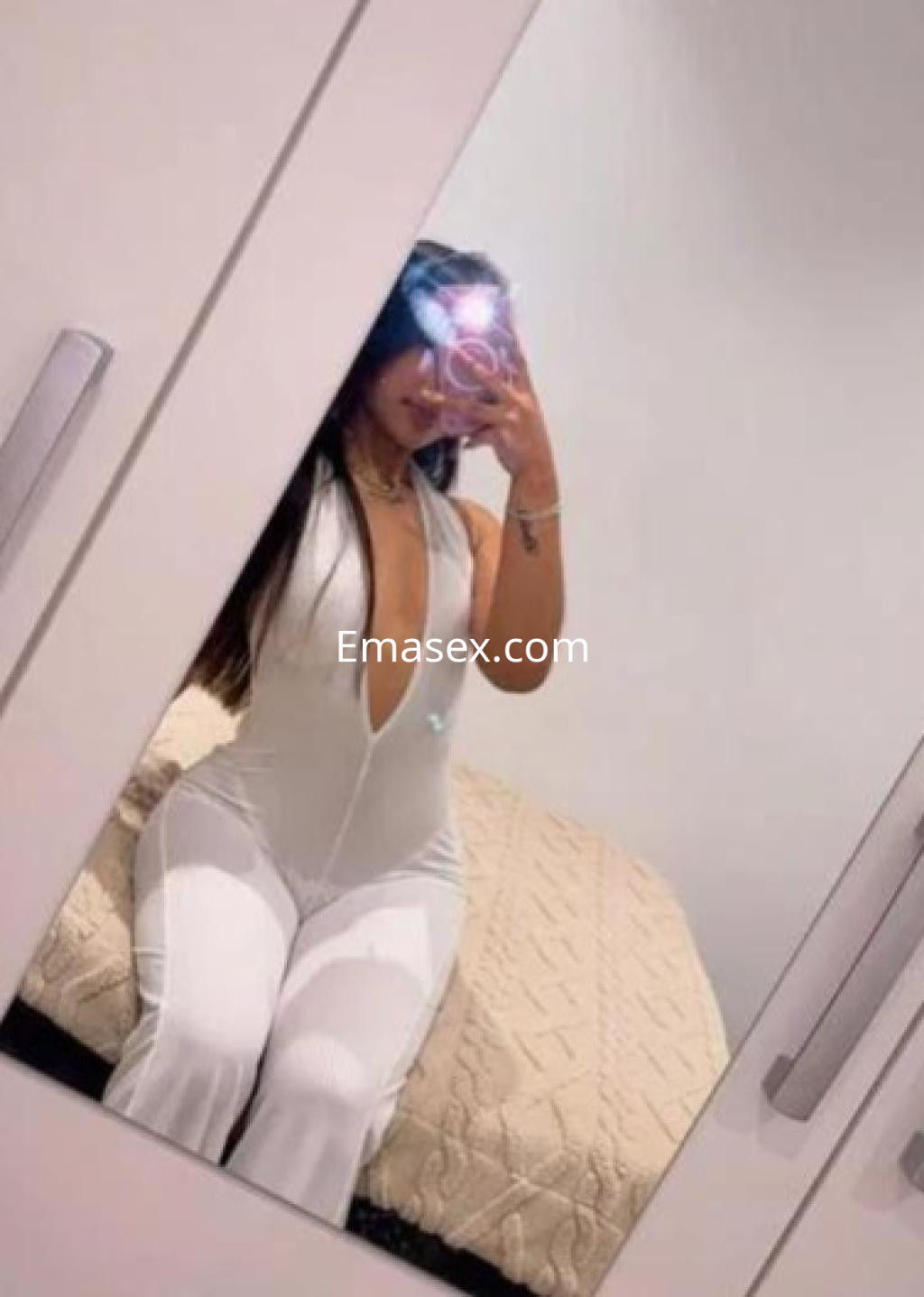 La puta se fotografía en el espejo, vistiendo un mono blanco con un profundo escote en una habitación de Murcia.