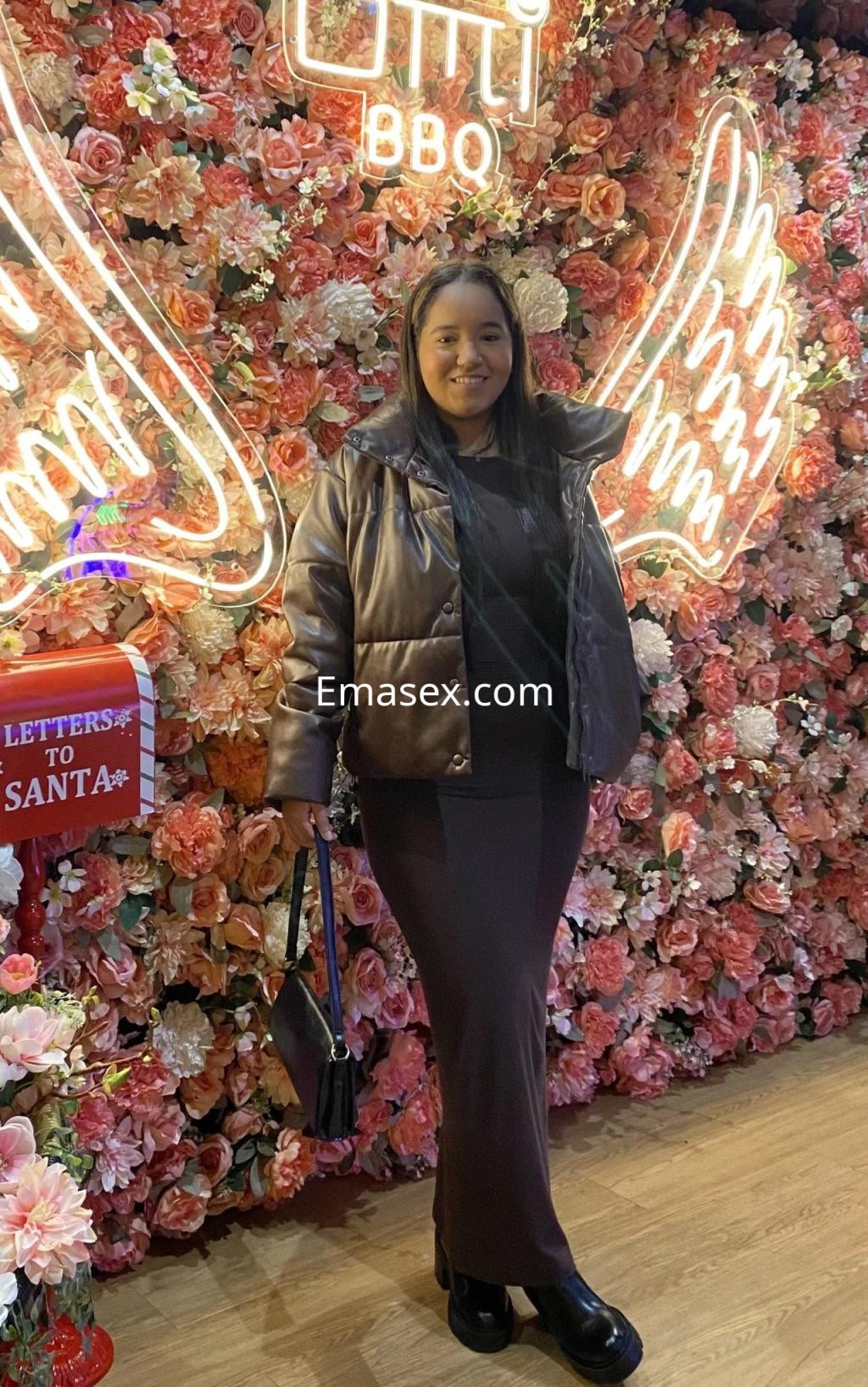 Escort en Vigo, de pie frente a una pared de flores, viste una chaqueta marrón y una falda larga negra.