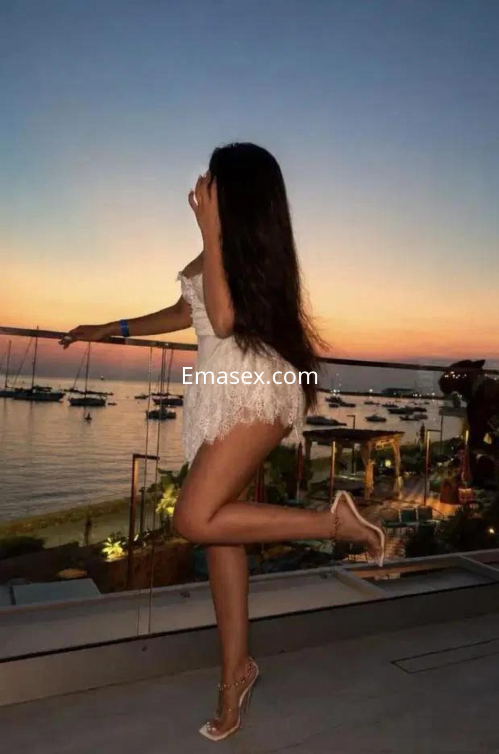 Escort en Almería, luciendo un vestido blanco y tacones, posando con el atardecer y el mar de fondo.