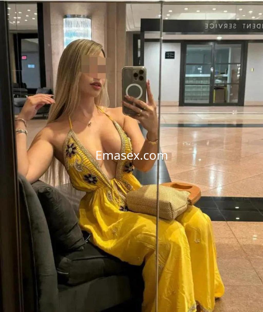 Puta en Almería posando en un vestido amarillo con escote, sentada en un sofá frente a un espejo.