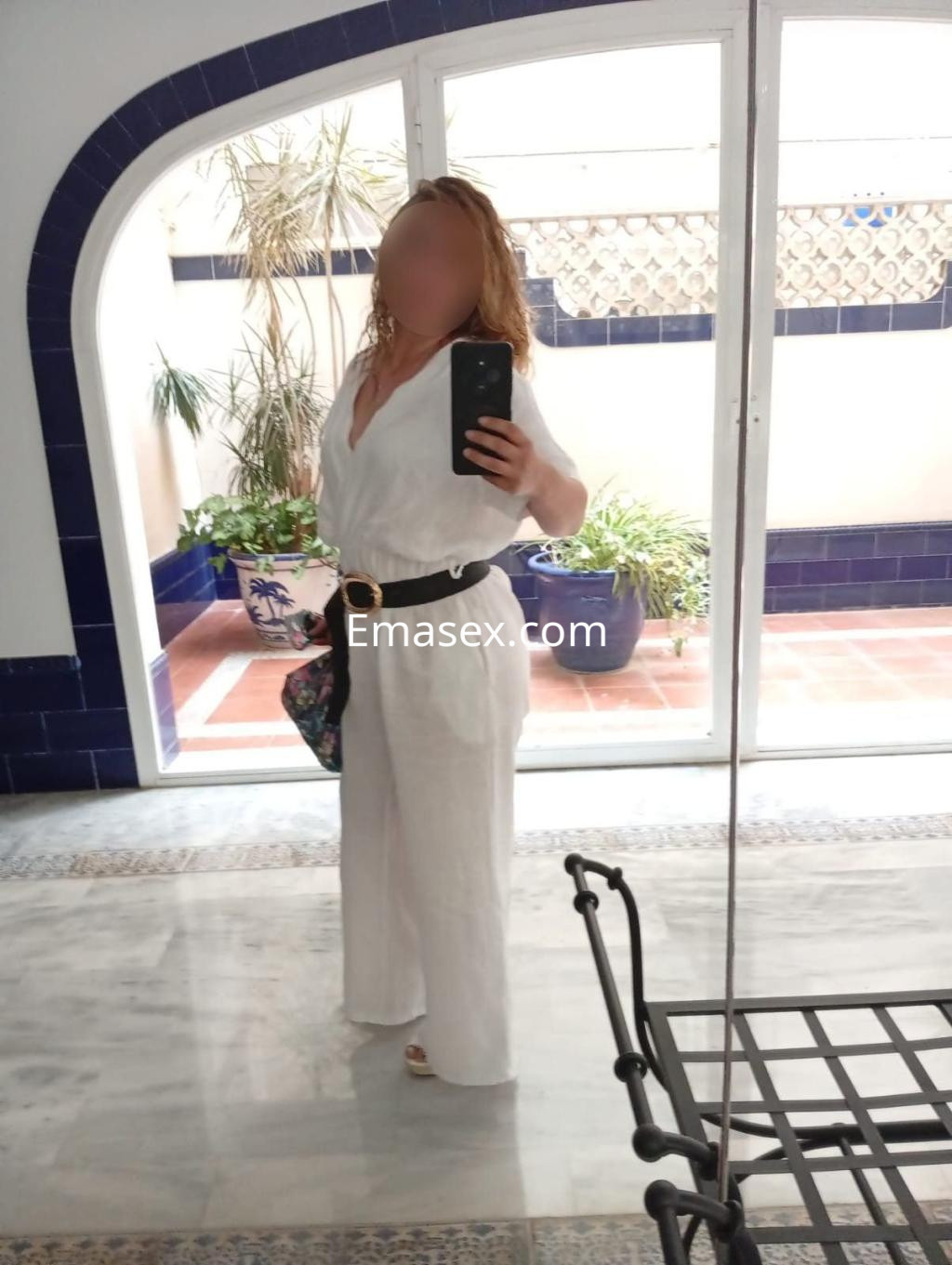 La puta en Málaga luce un mono blanco con un cinturón negro, posando frente a un espejo en un ambiente luminoso.