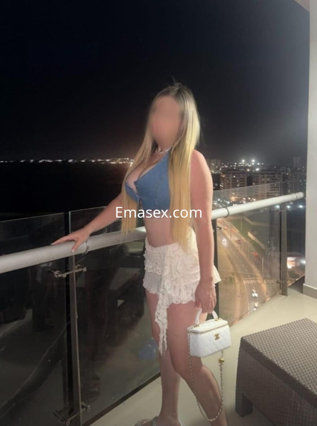 Escort en Granada con cabello largo y rubio, viste un top de mezclilla y una falda blanca, posando en un balcón.