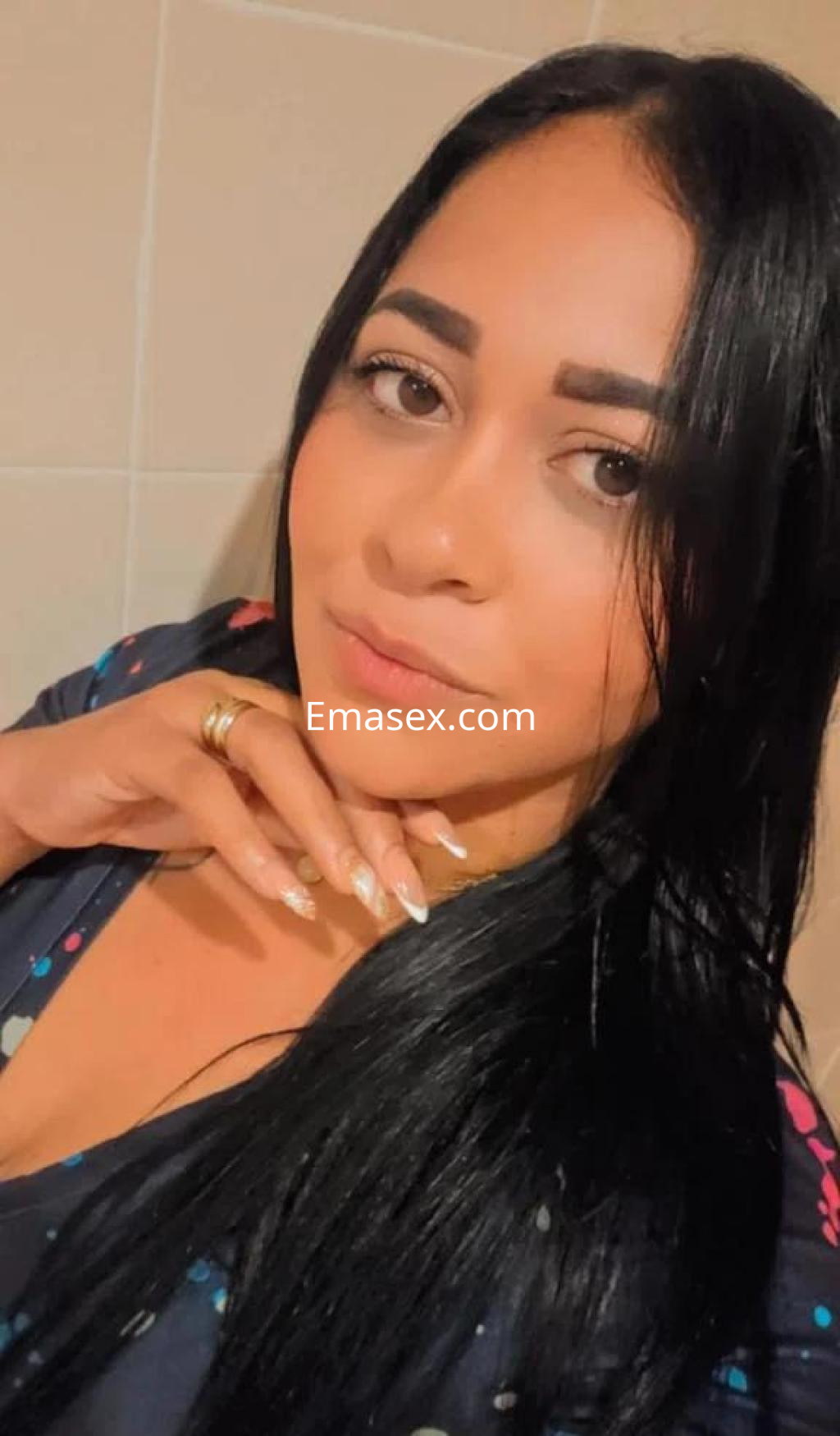 Escort en Madrid con cabello lacio y oscuro, viste una blusa con estampado colorido y posa con una mano en la cara.
