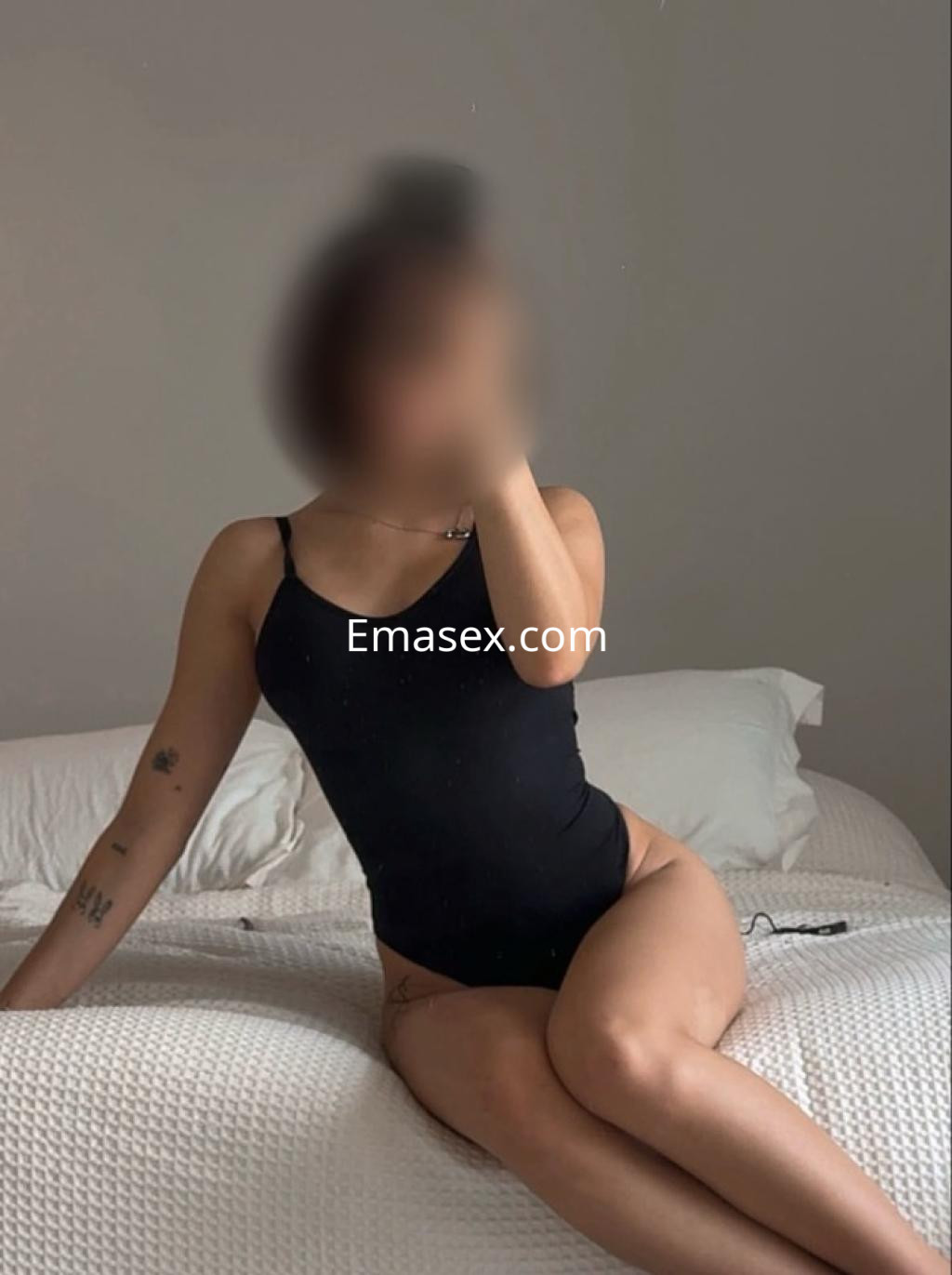 Escort sentada en la cama, vistiendo un bañador negro, con el pelo recogido y un fondo neutro en Tarragona.
