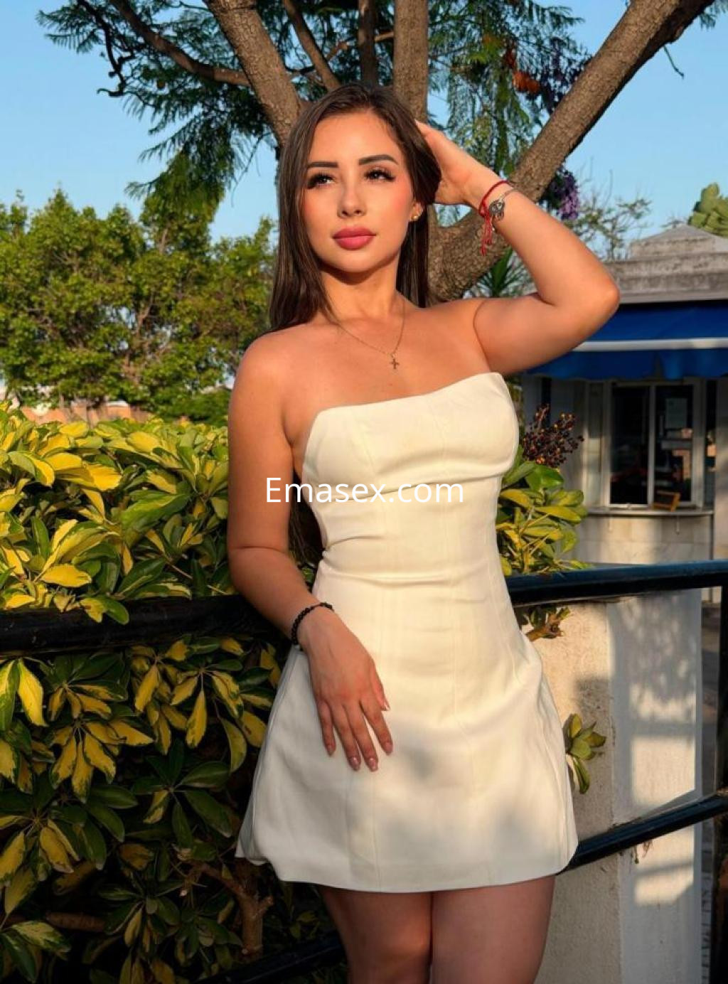 Puta en Málaga, luciendo un vestido blanco sin mangas, posando junto a una planta y un fondo natural.