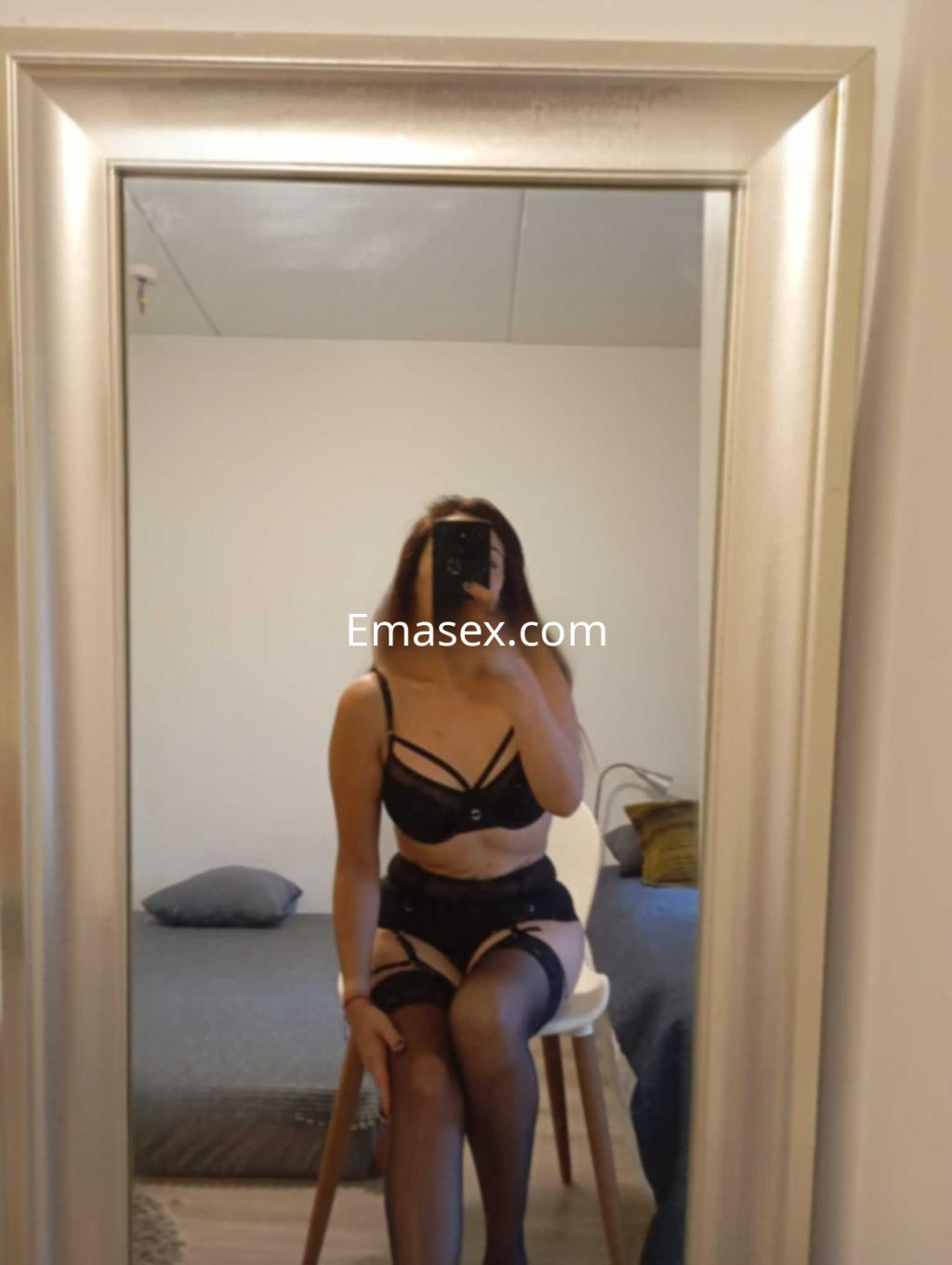 Escort en Valencia sentada en una silla, luciendo lencería negra con tirantes y medias, reflejada en un espejo.