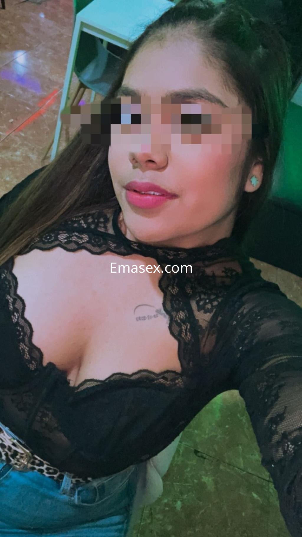 La puta posa con una blusa negra de encaje y un cinturón plateado, mostrando su escote en un ambiente iluminado.