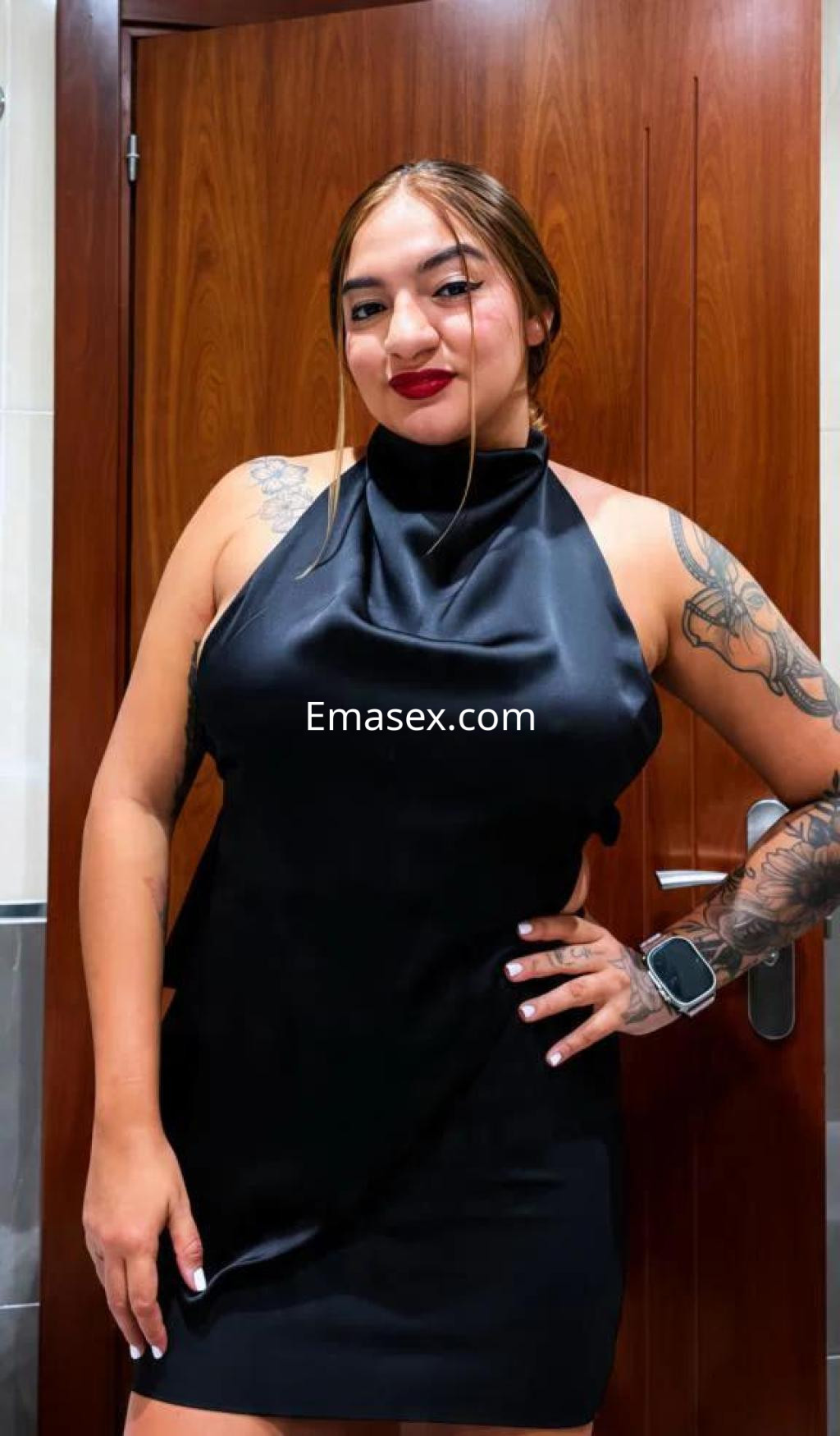 Escort en Lugo con vestido negro satinado, cabello recogido y tatuajes visibles en los brazos.