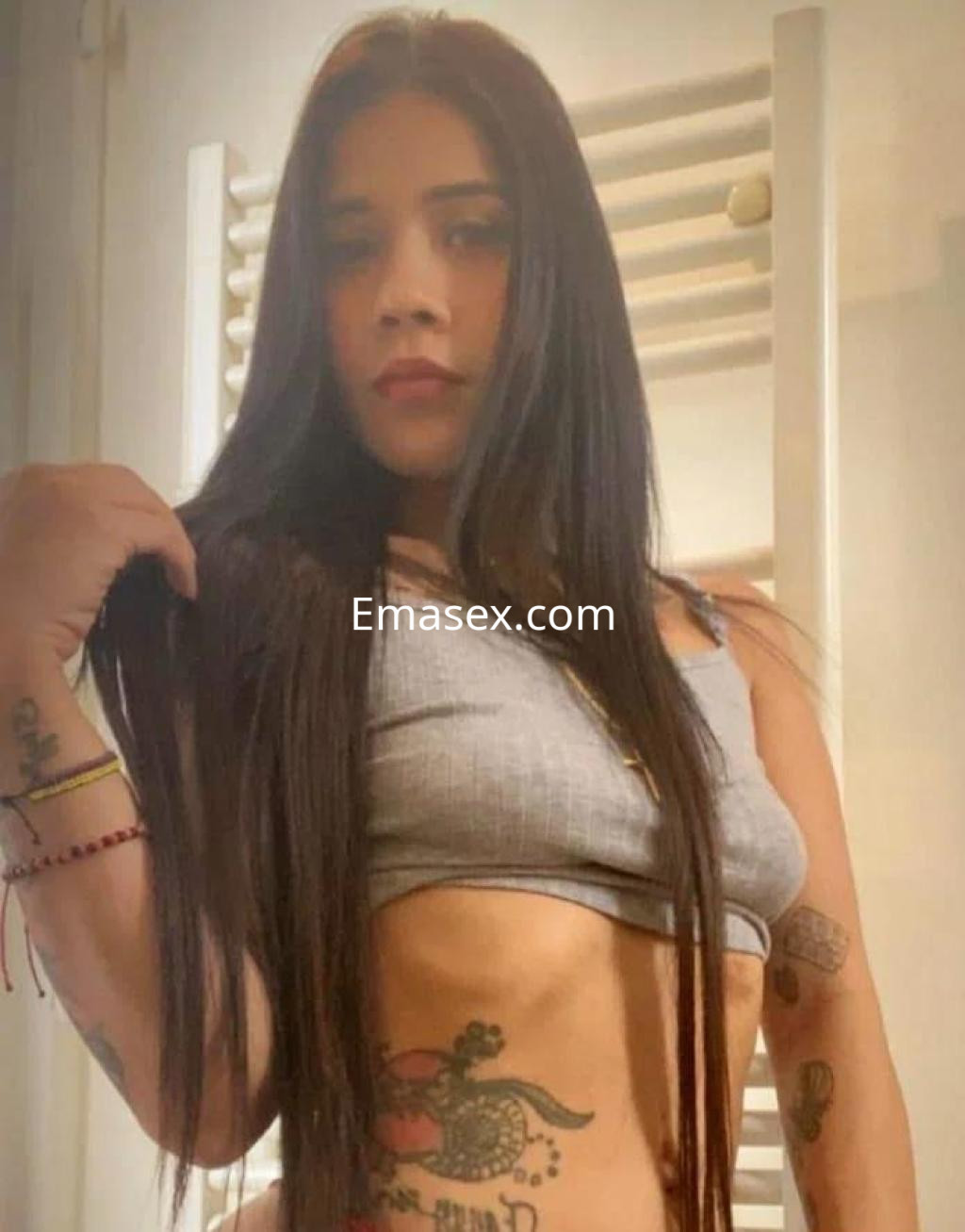 Puta en Alicante con cabello largo y oscuro, vistiendo un top gris que muestra su abdomen y tatuajes.