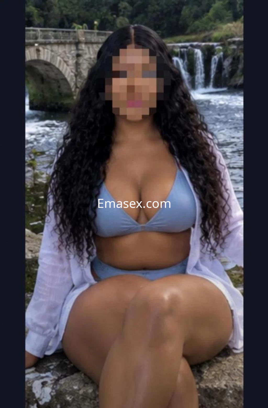 Puta posando a orillas de un río en Lugo, lleva un bikini azul y una camisa blanca abierta, con cabello rizado.