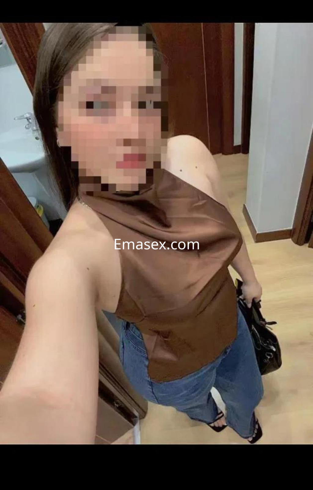 Escort en Zamora posando en un entorno interior, vistiendo una blusa marrón y jeans, con un bolso negro.