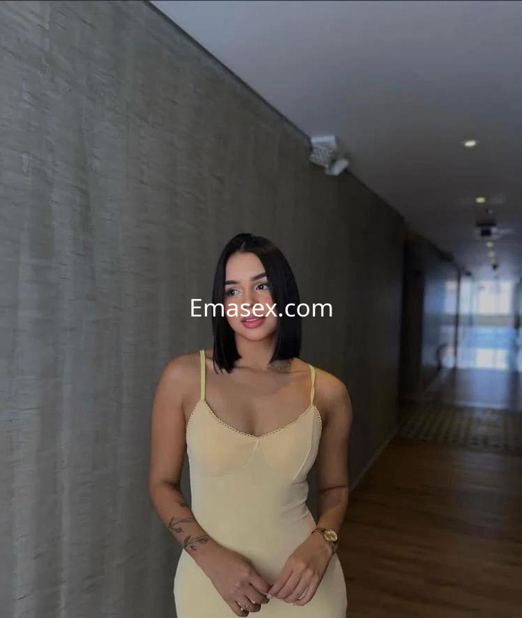 Escort en Palma de Mallorca, con vestido ajustado color nude y cabello negro, posando en un pasillo moderno.
