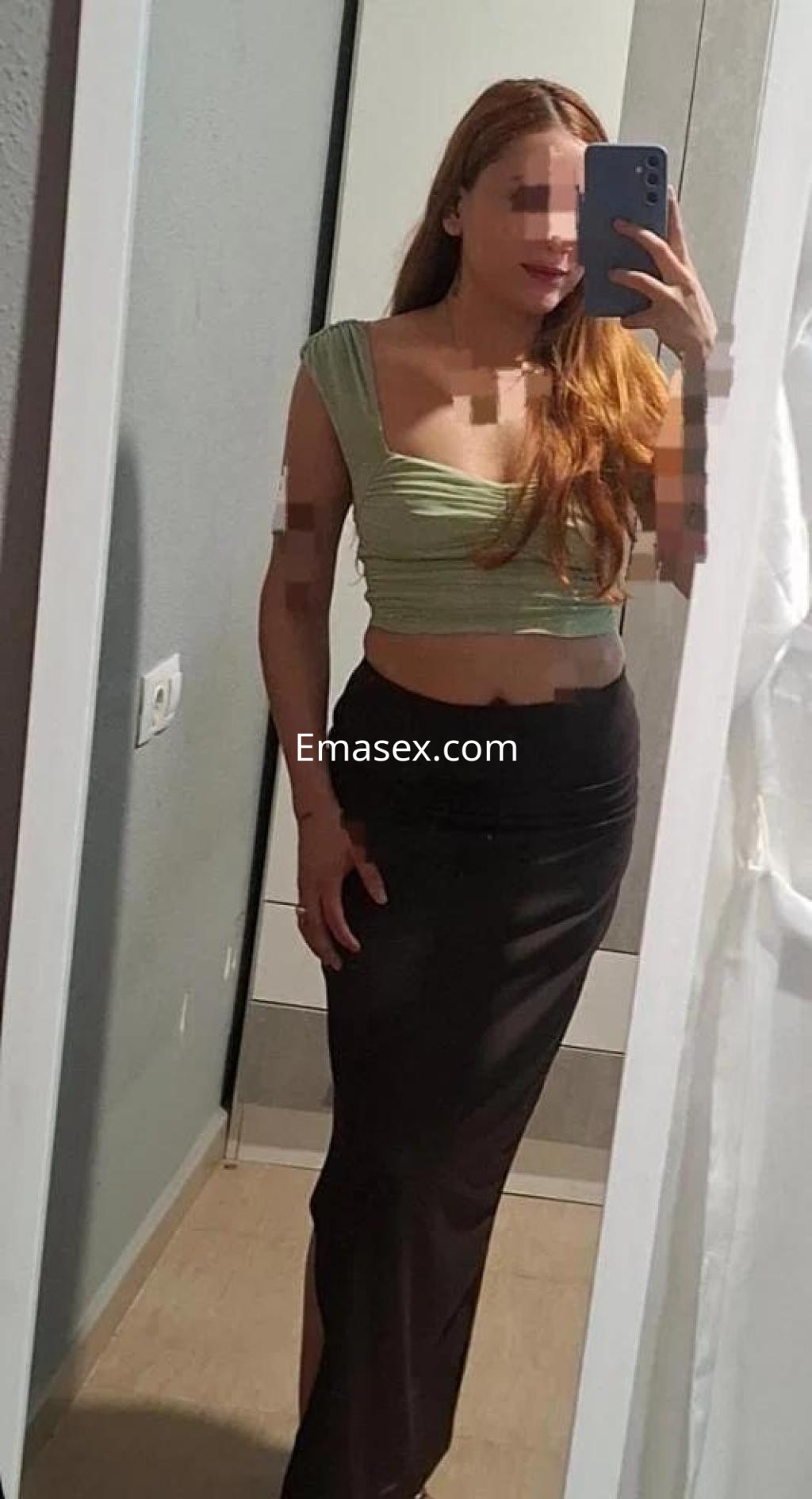 La puta en Alicante viste un top verde de tirantes y una falda larga negra, posando frente al espejo.