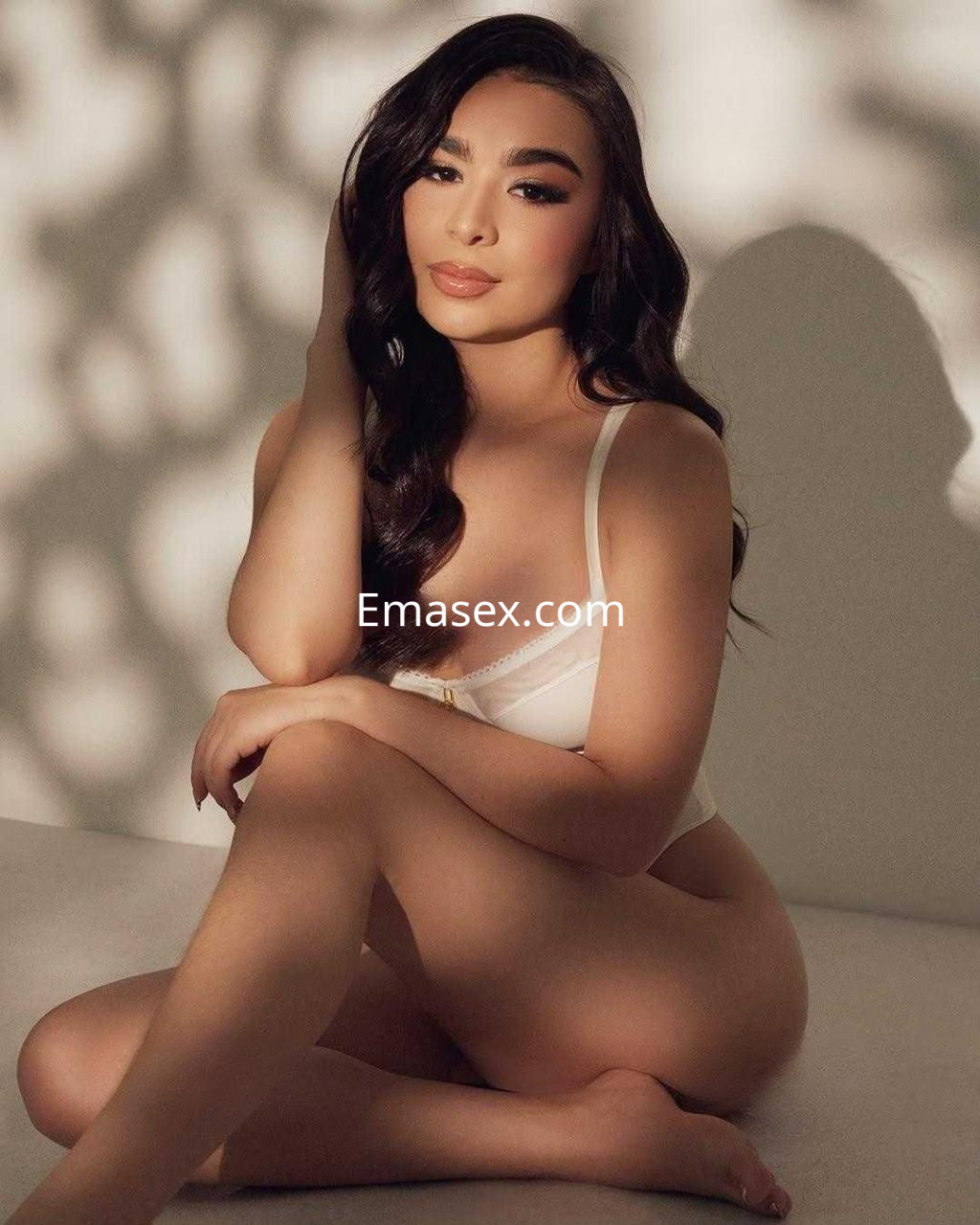 Escort en Barcelona sentada en una superficie clara, con lencería blanca y cabello ondulado, mostrando una expresión suave.