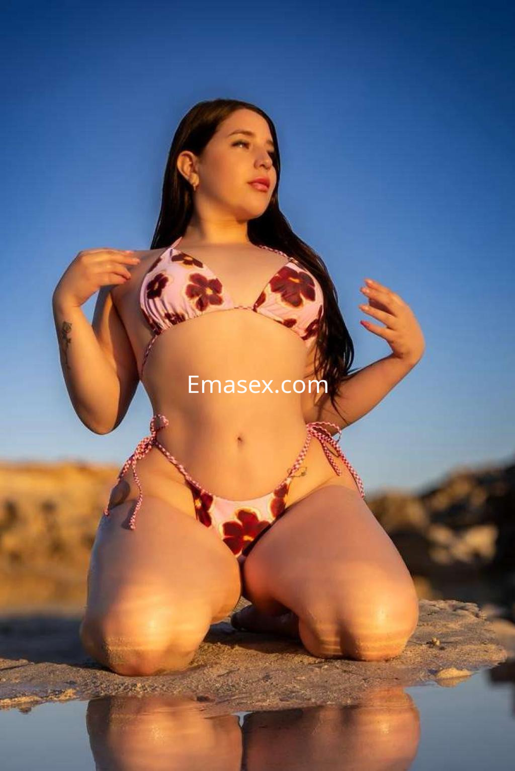 Escort en bikini floral posando de rodillas en la playa de Ibiza, con el sol brillando al fondo.